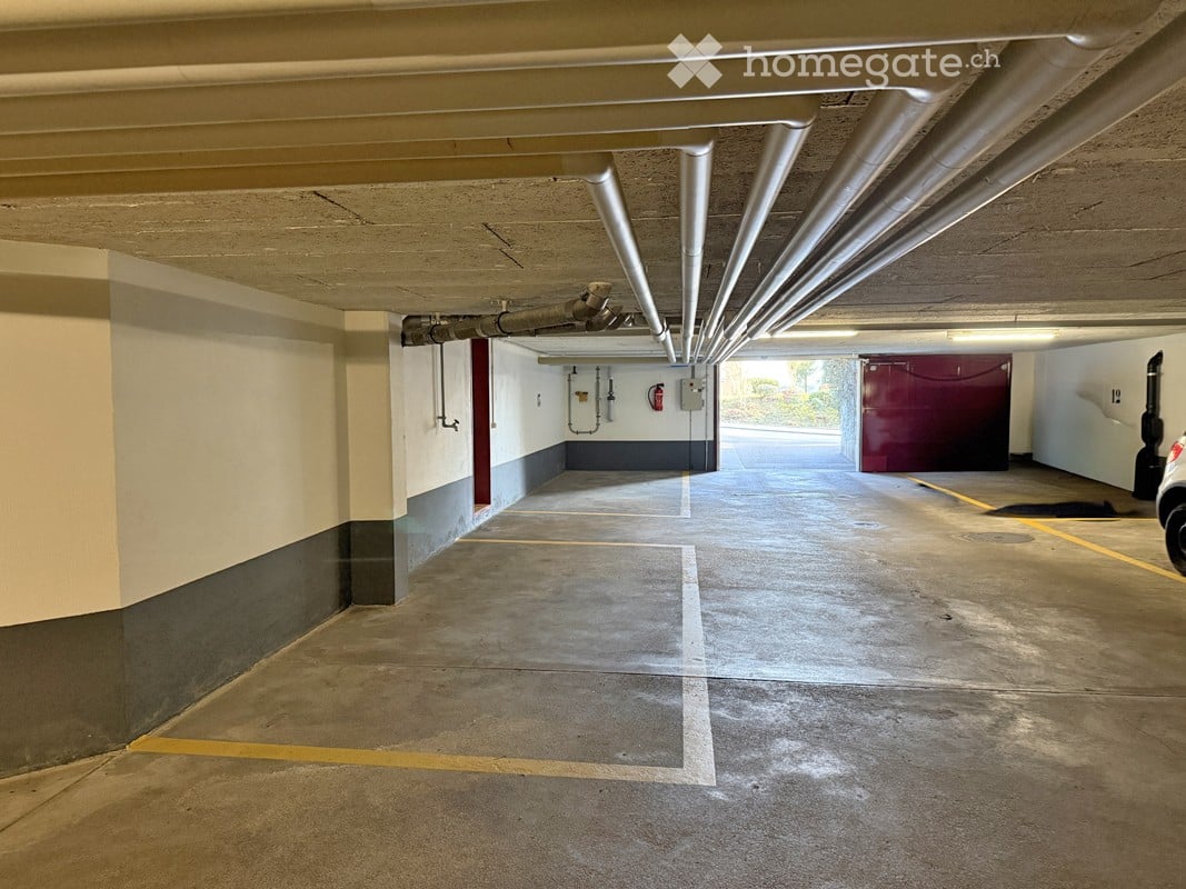Underground parking garage with marked parking spaces, concrete floors, and exposed pipes. A red storage unit is visible in the background.