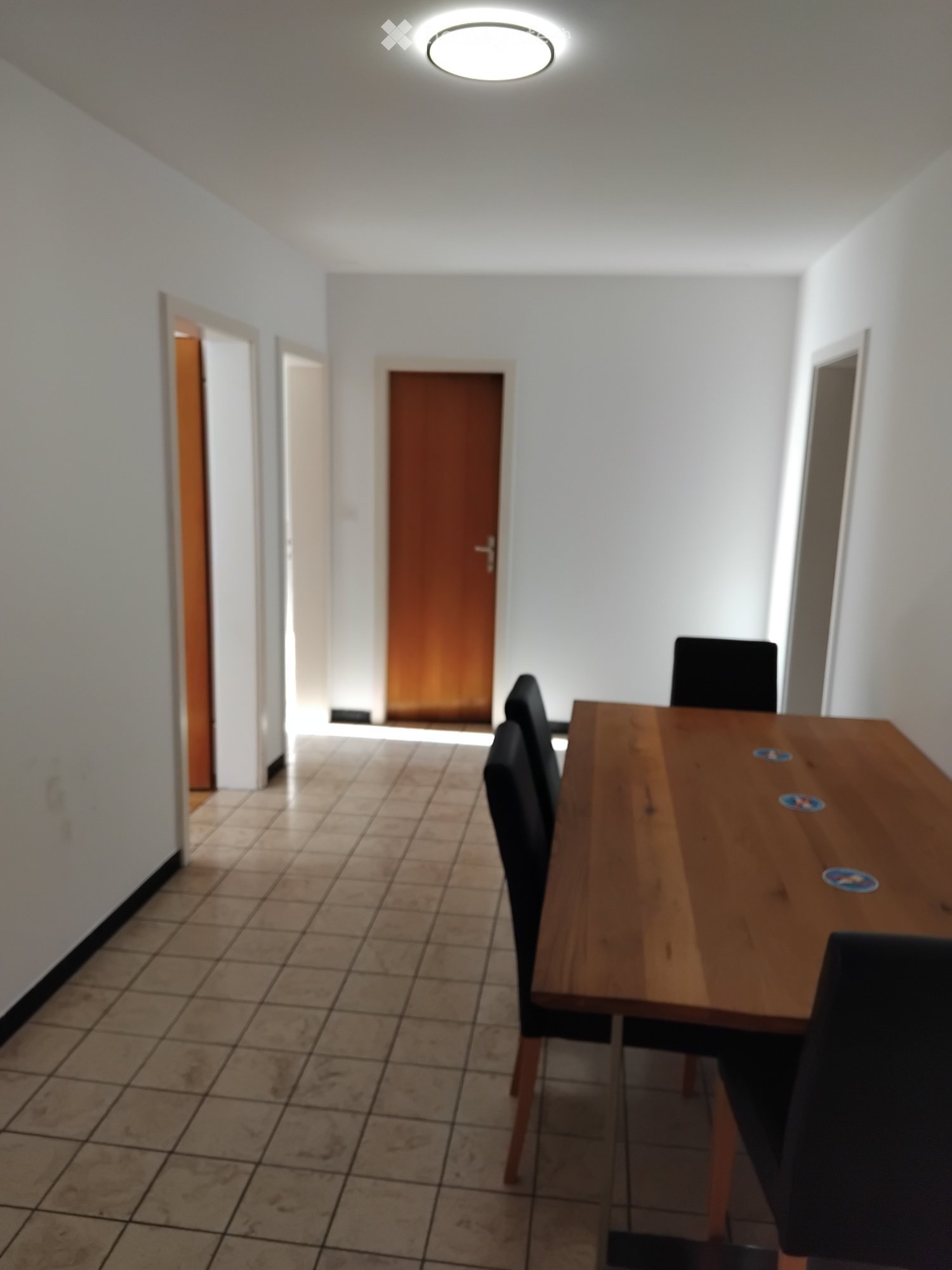 Dining area with a wooden table and black chairs, tiled floor, white walls, and recessed ceiling light.
