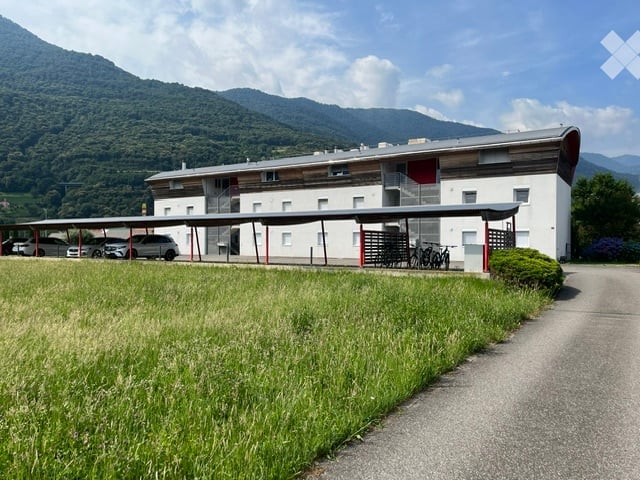 Modern multi-story building with a covered parking area, surrounded by a grassy field and mountain backdrop.
