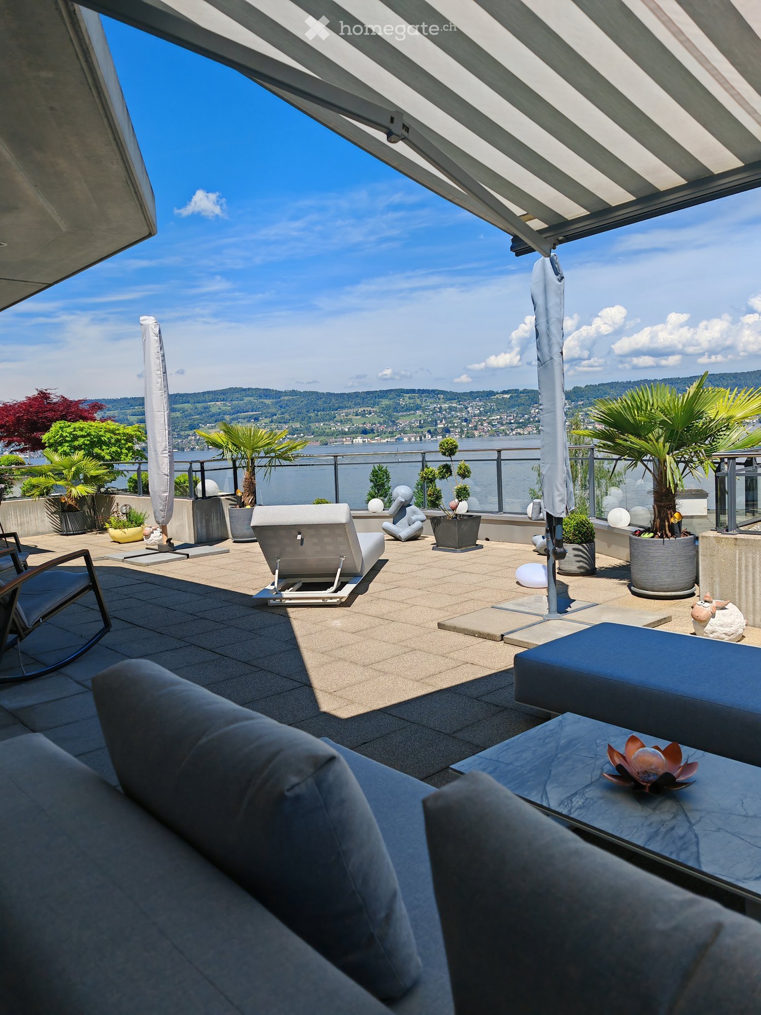 Modern rooftop terrace with panoramic view, featuring a sofa, chairs, a table, and potted plants.