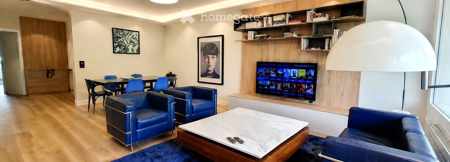 Modern living room with blue leather sofas, a marble coffee table, a TV, a dining area with chairs and a table, and built-in shelving with books and decor.