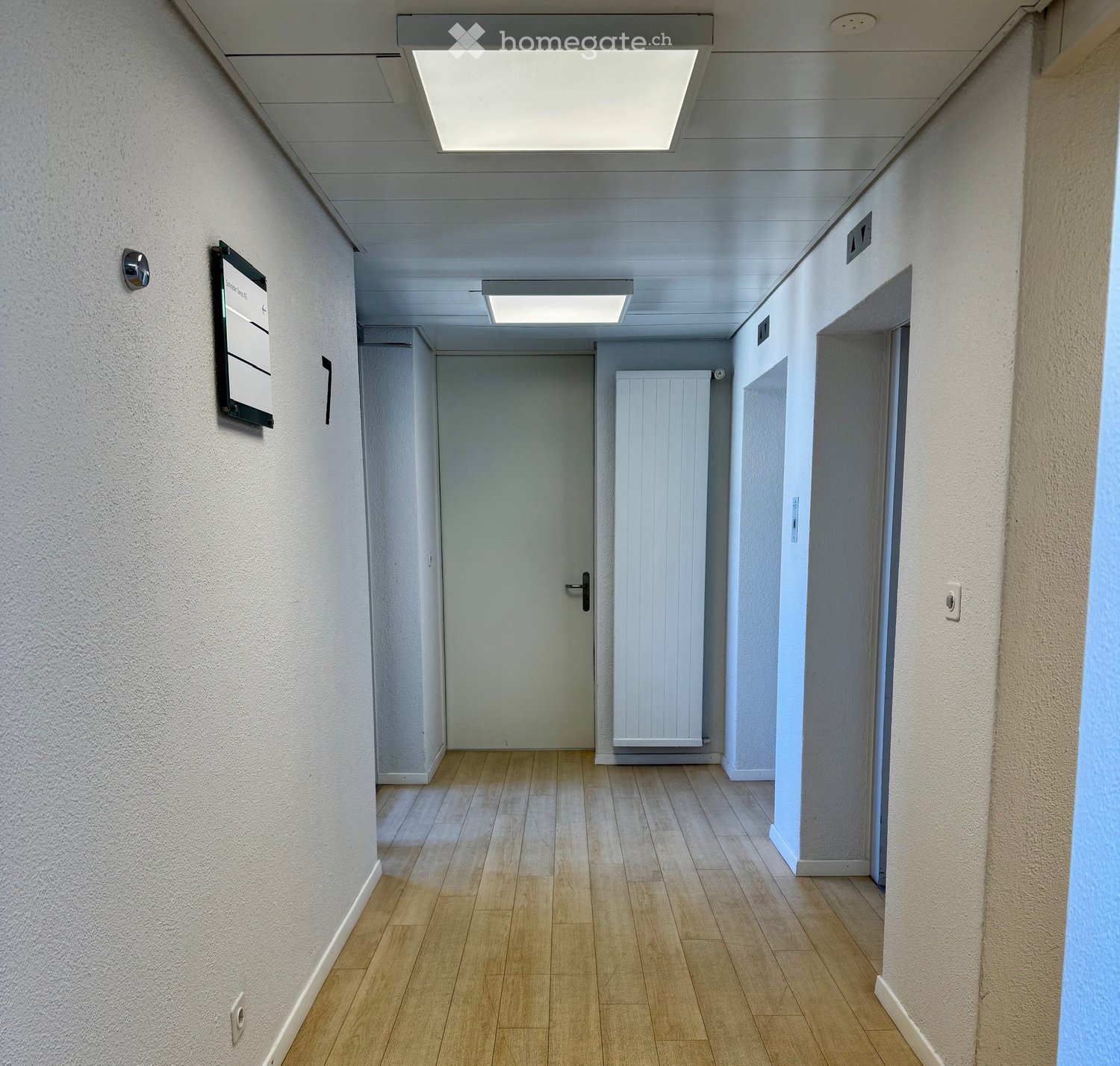 Clean, well-lit hallway with wooden flooring, white walls, and a closed door at the end. Features a radiator on the right wall and a signboard on the left wall.
