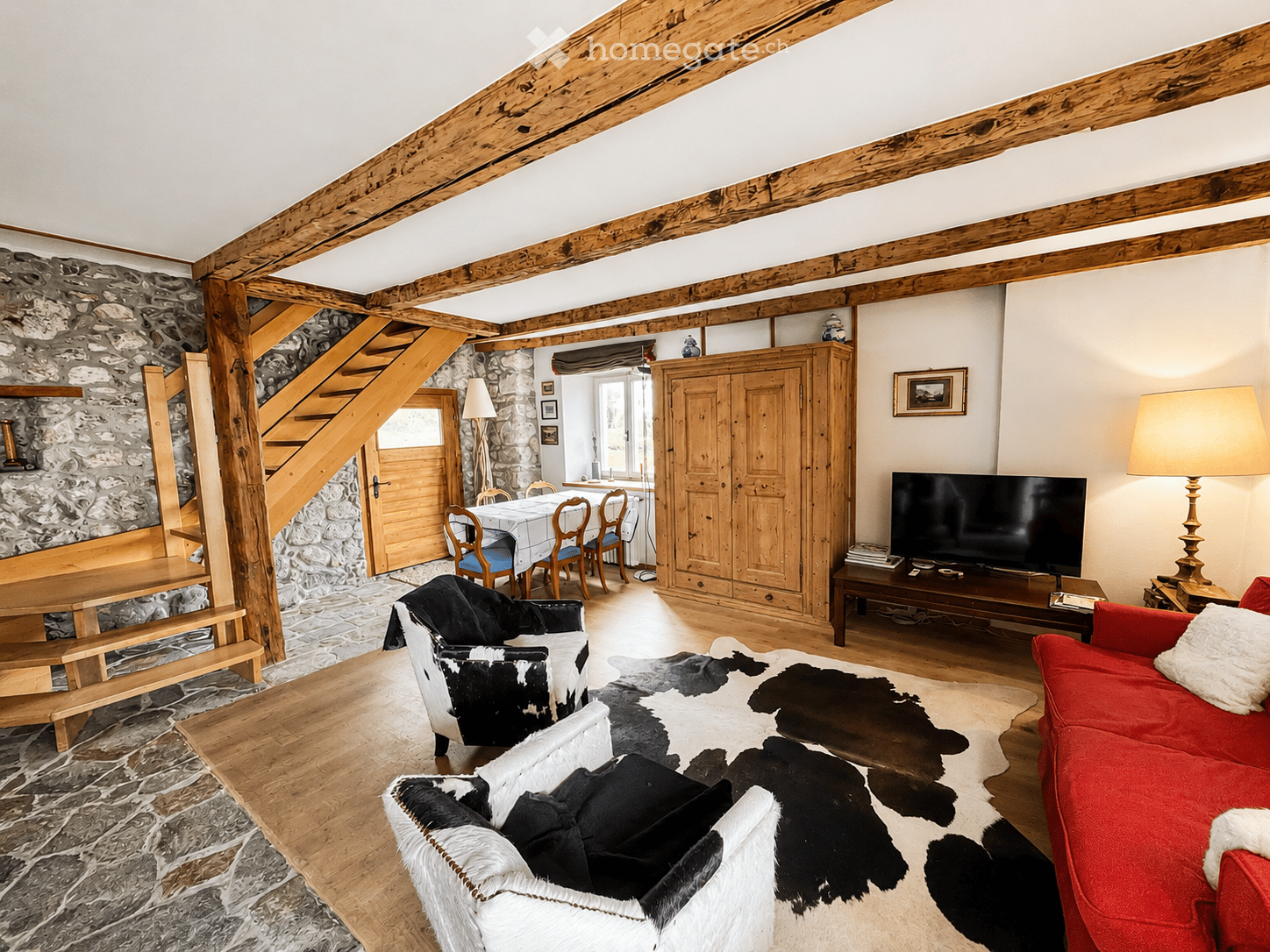 Cozy living room with exposed wooden beams, stone walls, a red sofa, a white cowhide rug, a coffee table, a TV, a dining area with a table and chairs, and a wooden cabinet.