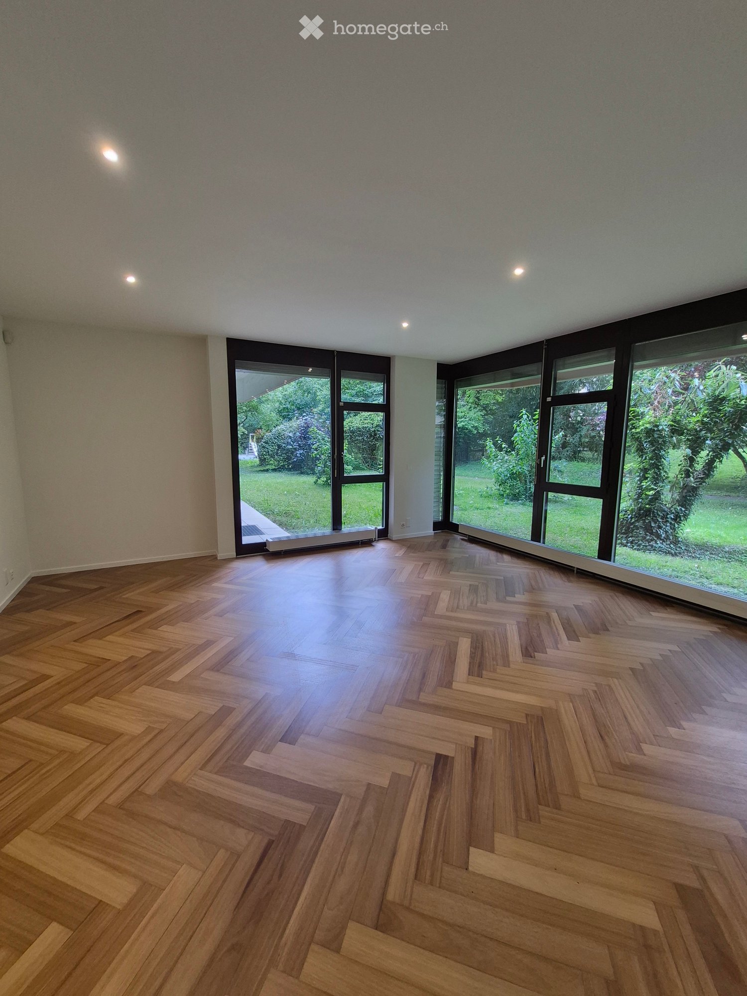 Empty room with wooden herringbone flooring, large windows, and recessed lighting.