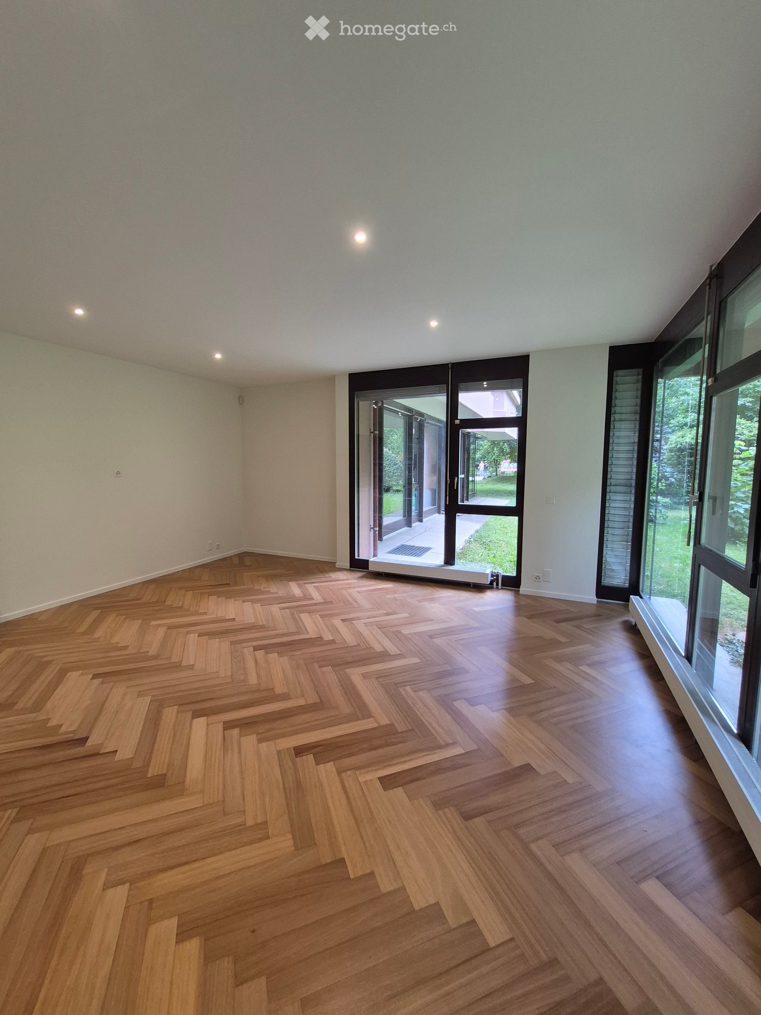 Empty room with herringbone-patterned wooden flooring, large windows, and a set of double doors leading outside.
