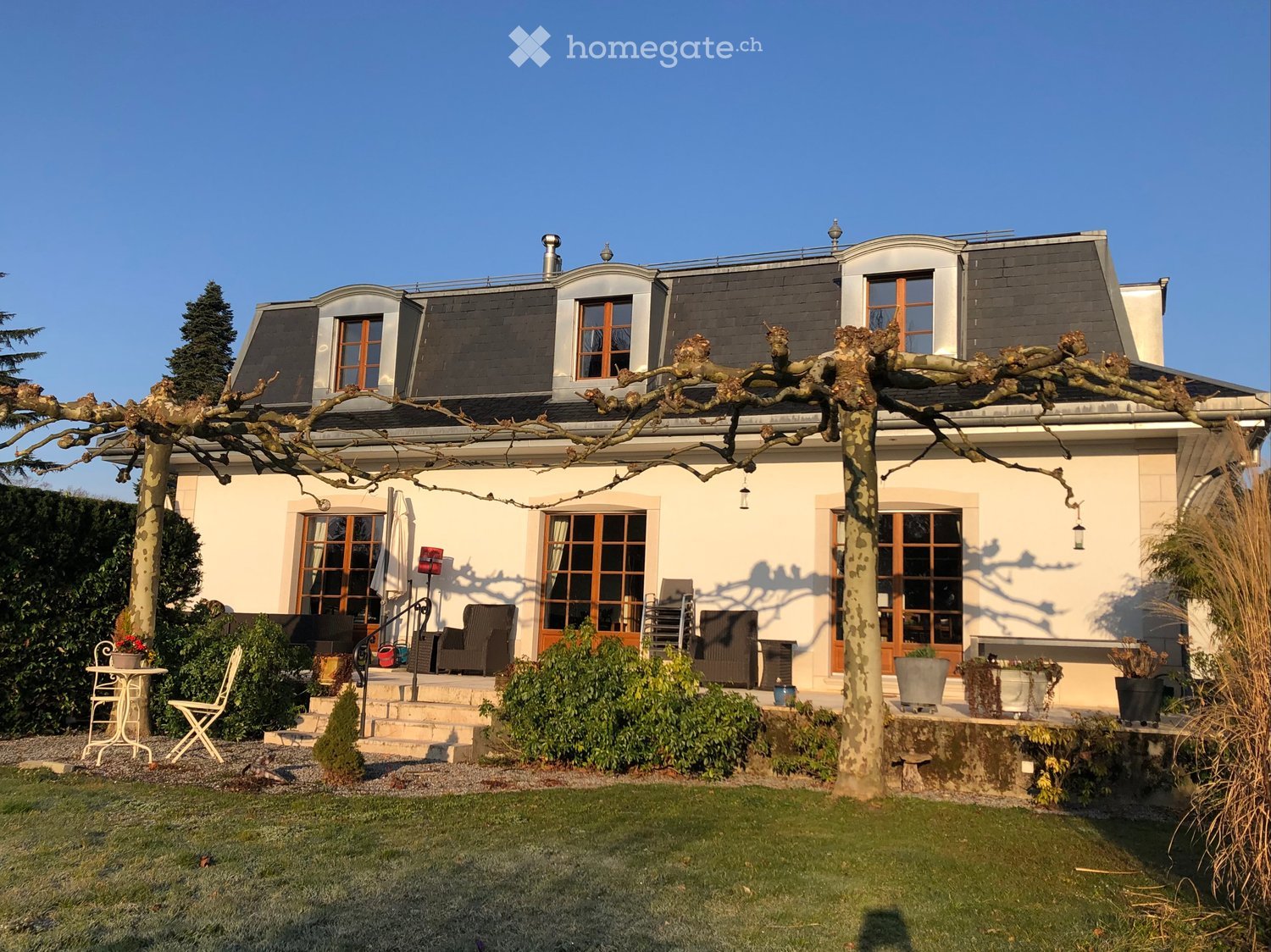 Two-story house with a gray roof, white walls, and large windows. The front features a well-maintained garden with a patio area, outdoor seating, and a pergola with climbing plants.