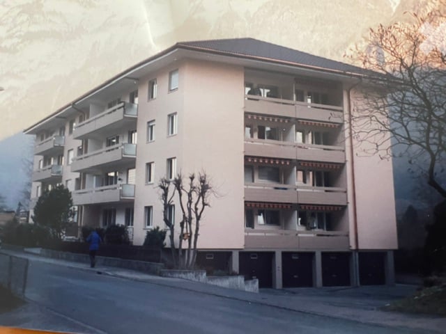Multi-story residential building with multiple windows and balconies, surrounded by trees and a road.