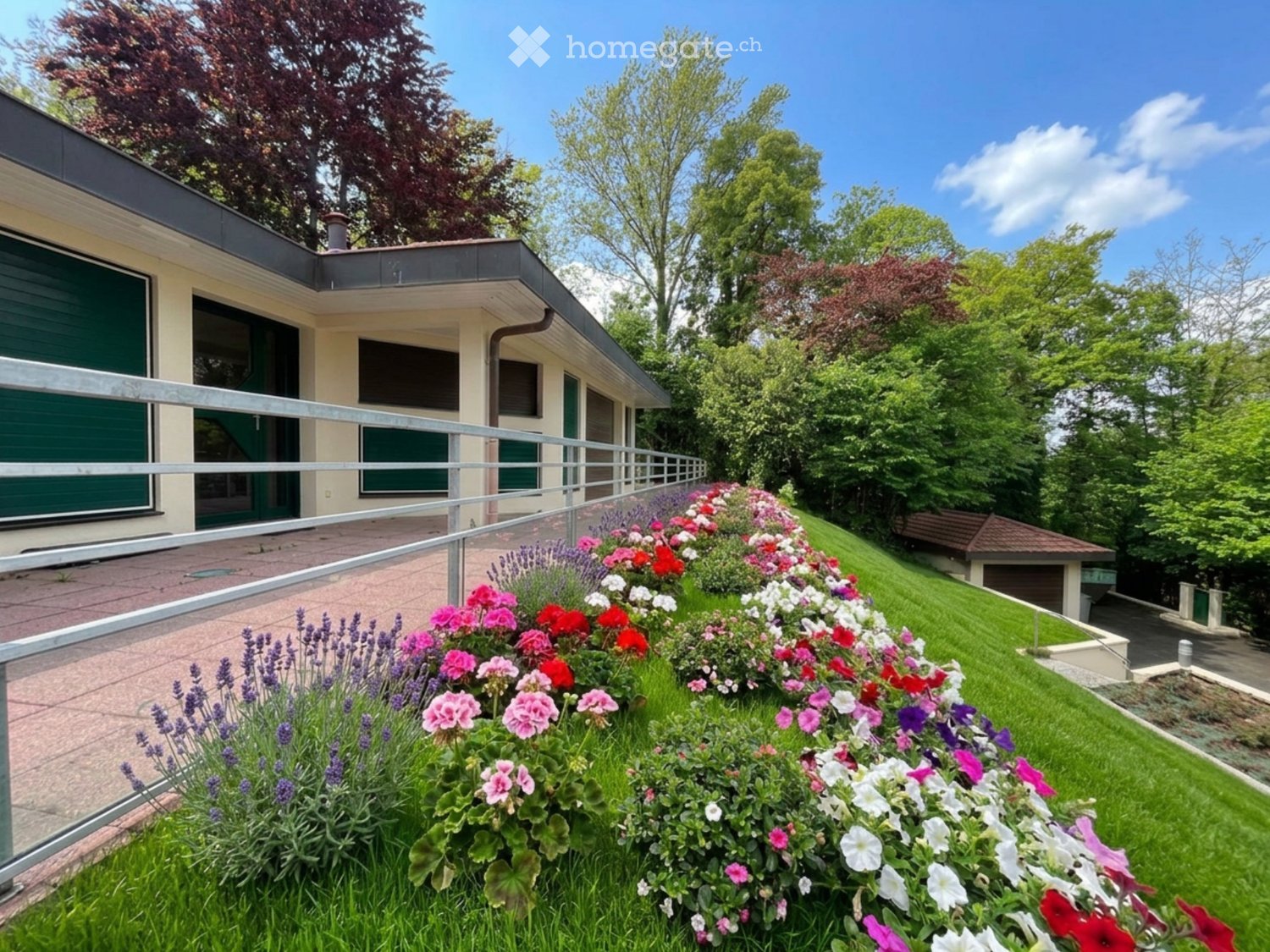Well-maintained garden with colorful flowers, lavender, and neatly trimmed grass, adjacent to a modern house with a balcony and garage.