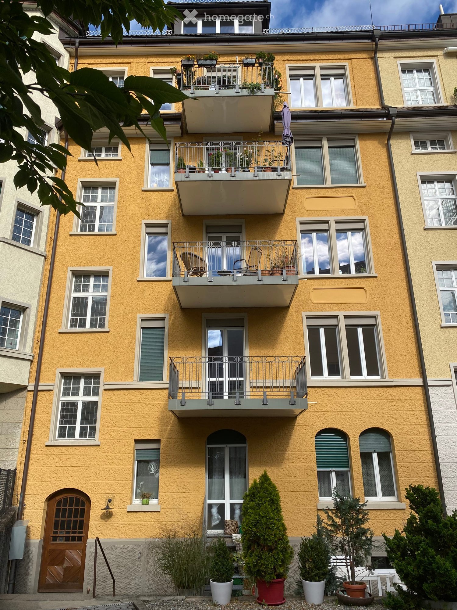 Four-story yellow building with multiple balconies, potted plants, and a wooden front door.