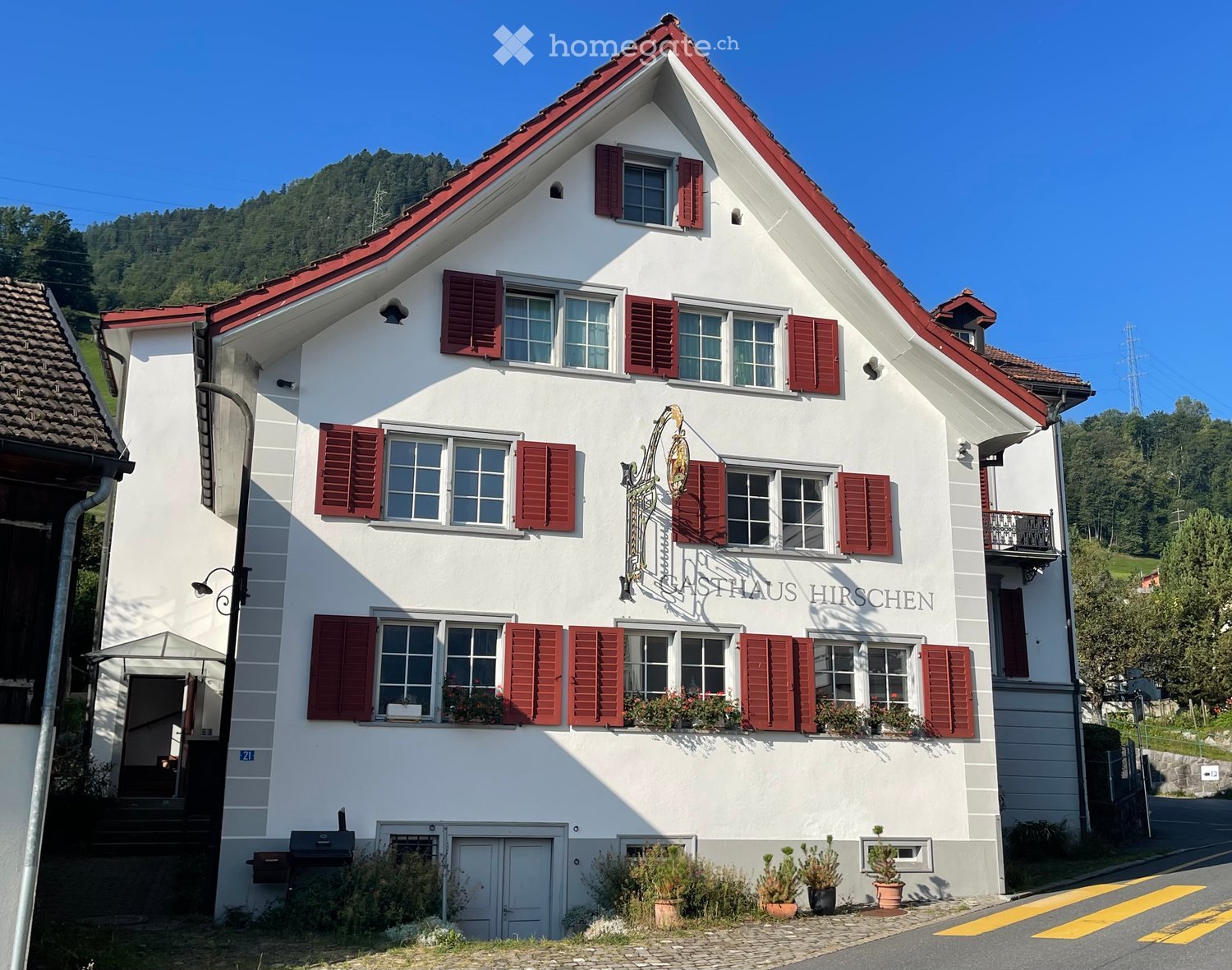 Two-story white house with red shutters and a red-tiled roof, featuring a sign reading "GASTHAUS HIRSCHEN" and small potted plants near the entrance.