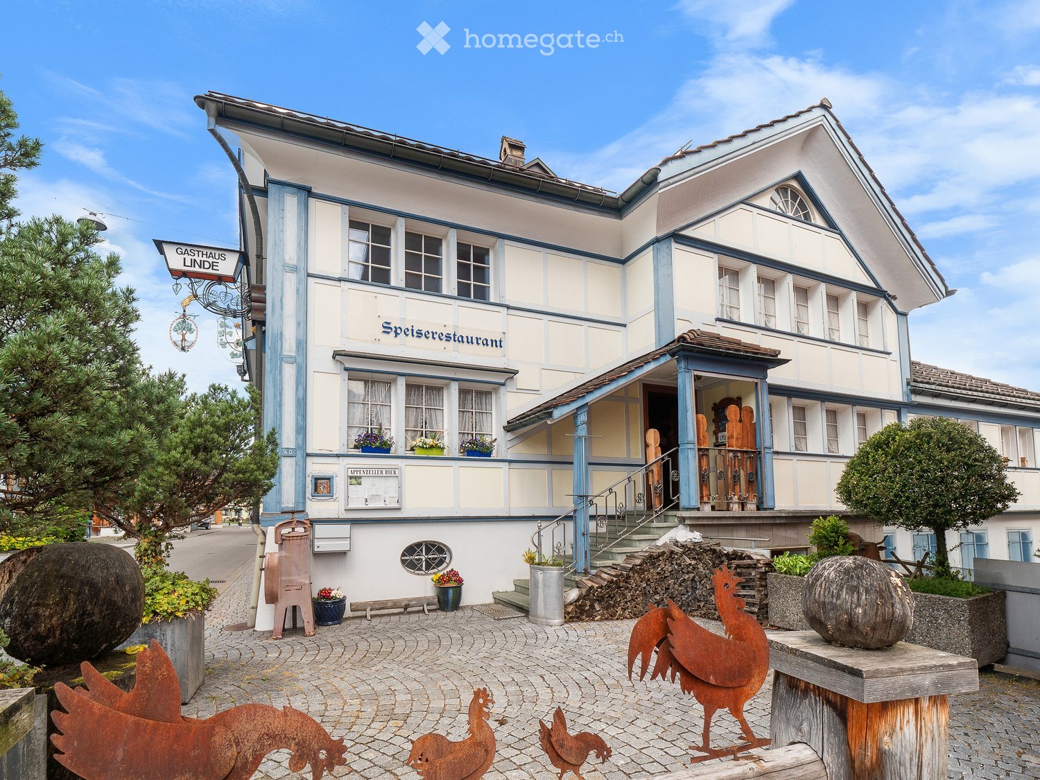 Two-story building with white and blue trim, labeled "Speiserestaurant" and "GastHaus Linde" with a balcony, decorative roosters, and potted plants.