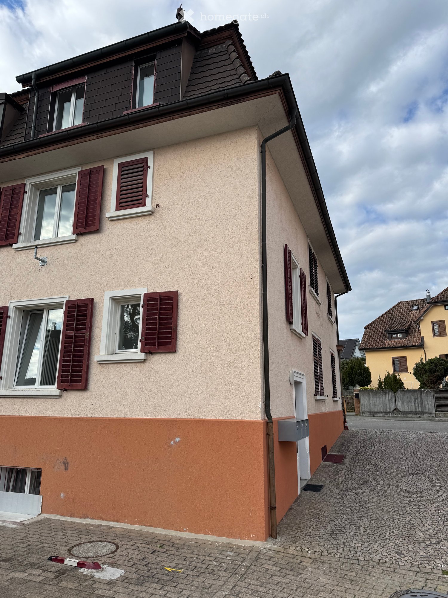 3 story house, light brown wall with red trims, brown roof, several windows, mailbox attached to wall