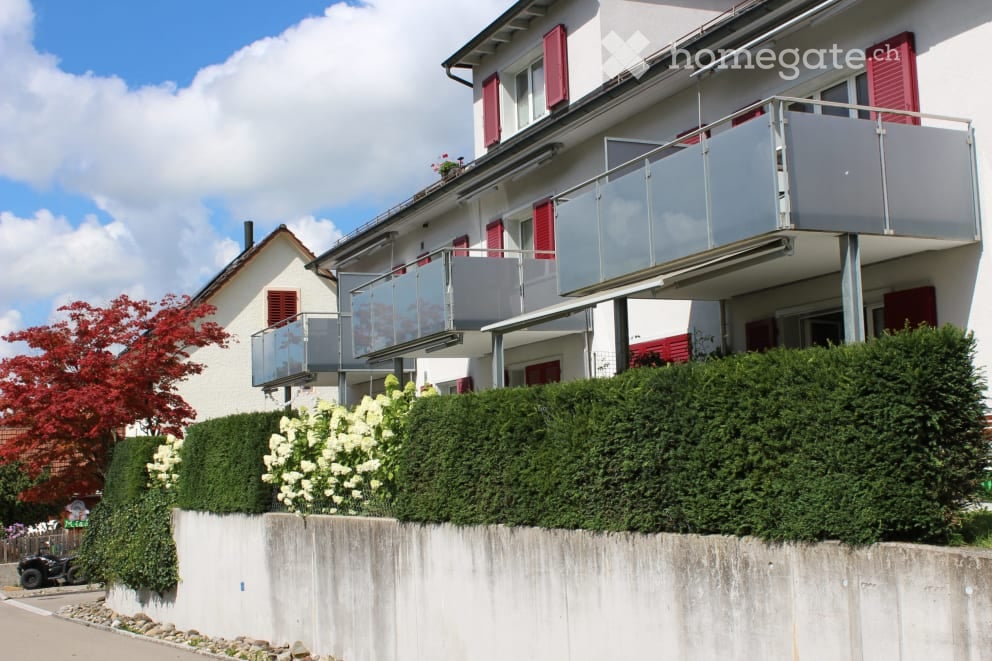 Two-story residential building with white walls, red shutters, and a large glass balcony overlooking a landscaped garden area.
