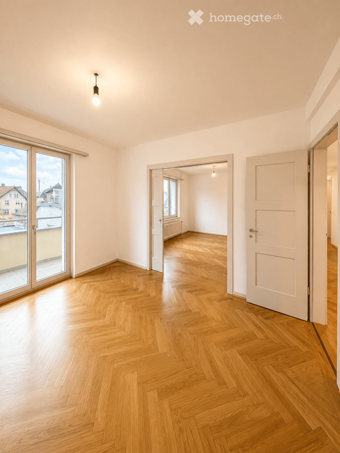 Empty room with wooden herringbone flooring, white walls, and a large sliding glass door leading to a balcony.