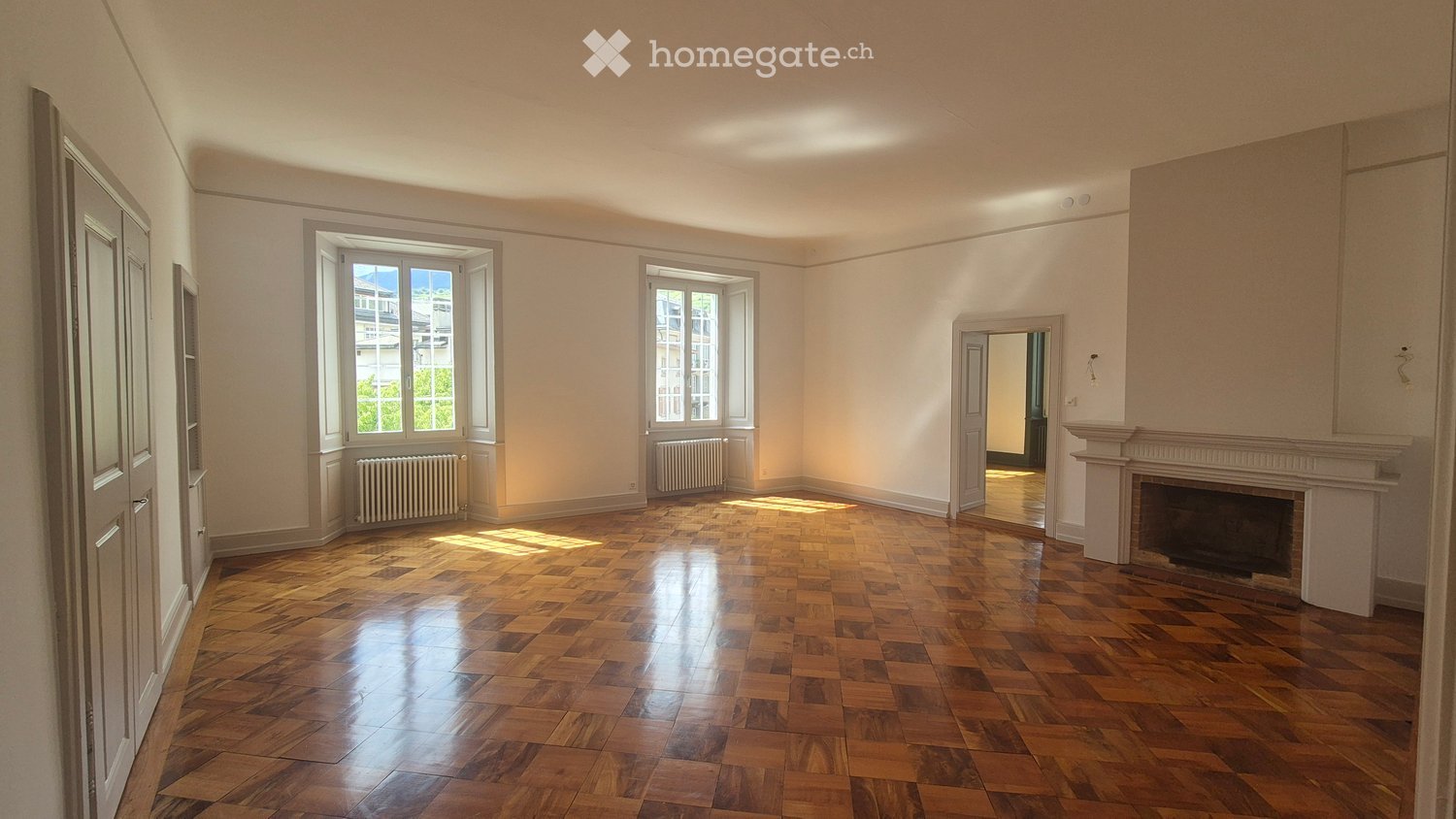 Empty room with wooden parquet flooring, white walls, large windows, and a fireplace. No furniture or equipment present.