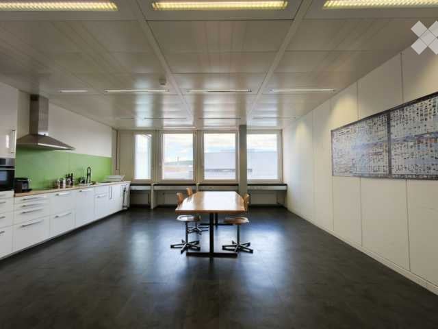 Bright, modern break room with a central table and chairs, kitchen area with cabinets, sink, and microwave, large windows providing natural light, and a colorful artwork on the wall.