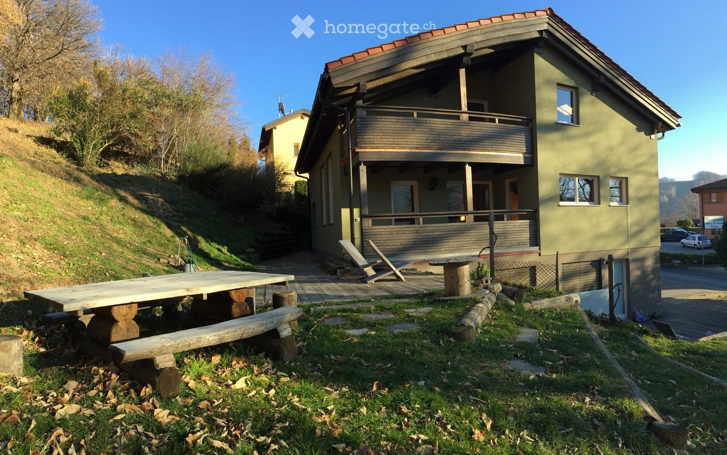 House. A modern house with a red roof and green walls, situated on a slope with a wooden deck and outdoor furniture.