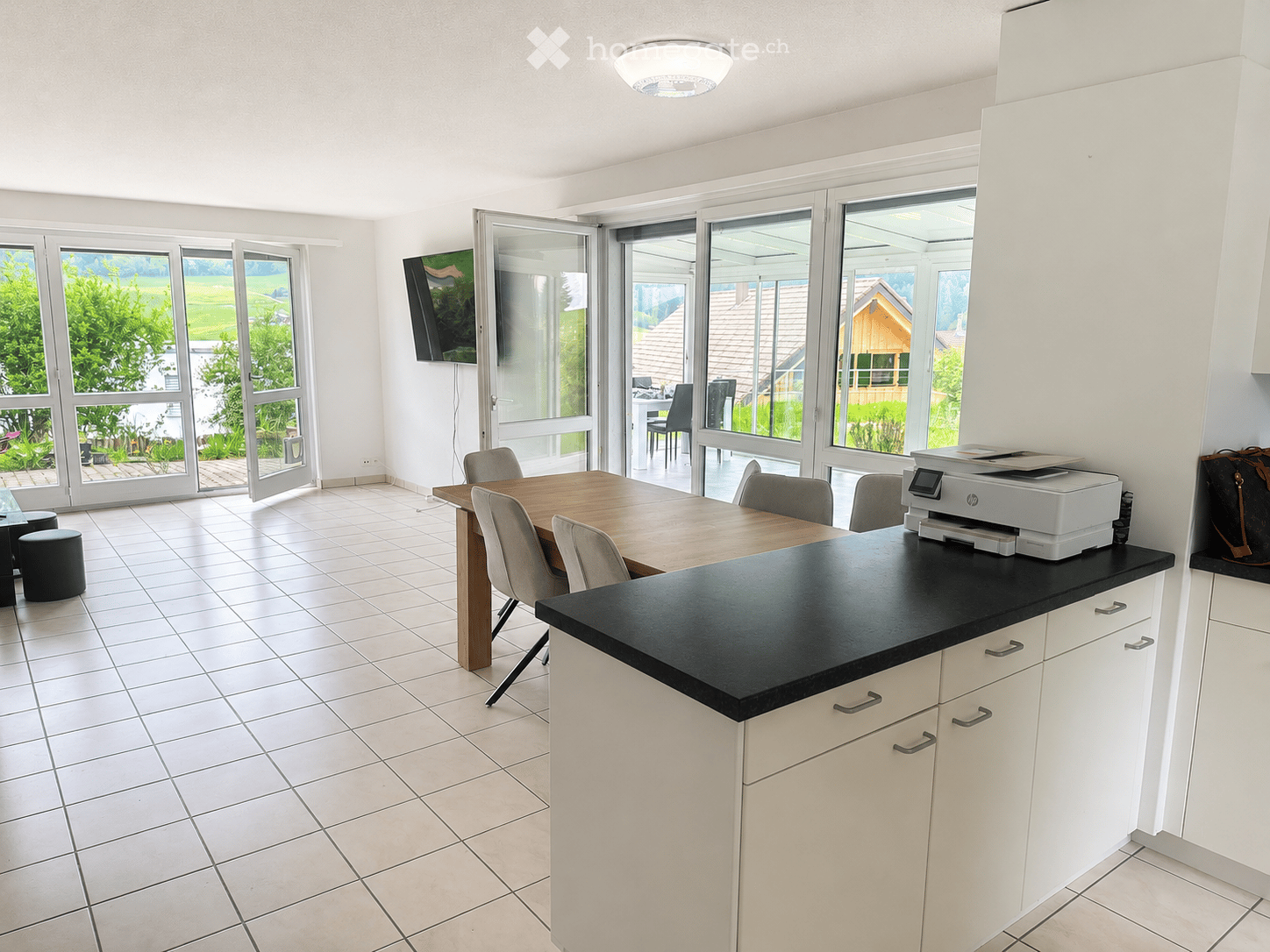 Bright kitchen with white cabinets, black countertop, tiled floor, and large windows overlooking a green landscape. Includes a dining table with chairs, a printer on the counter, and a TV mounted on the wall.