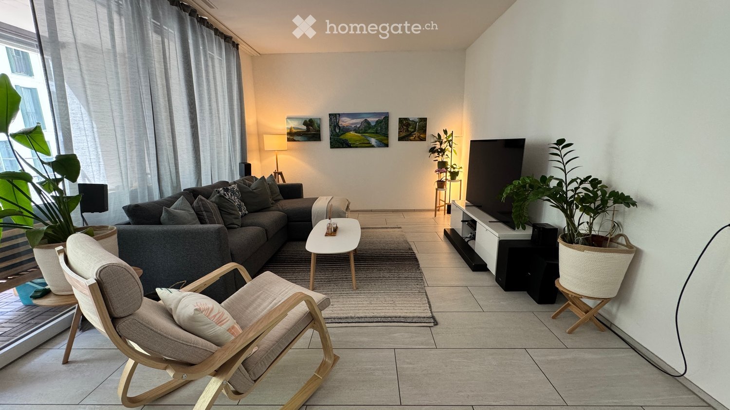 Modern living room with a grey sectional sofa, wooden coffee table, armchair, TV, and plants.