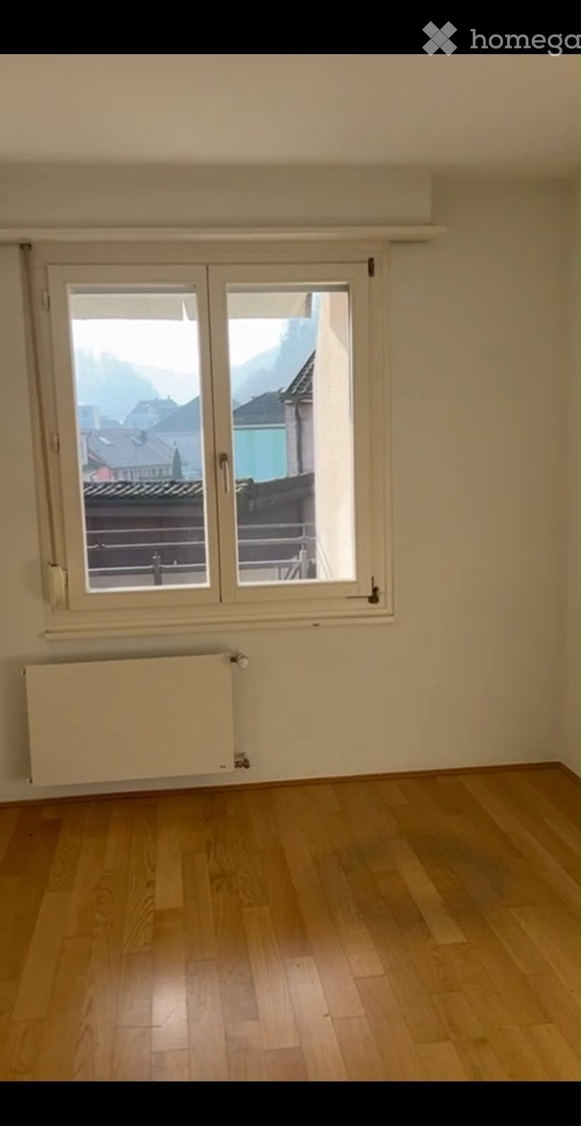 Empty room with wooden flooring, large window with double doors leading to a balcony, radiator below the window, and a view of rooftops and mountains.