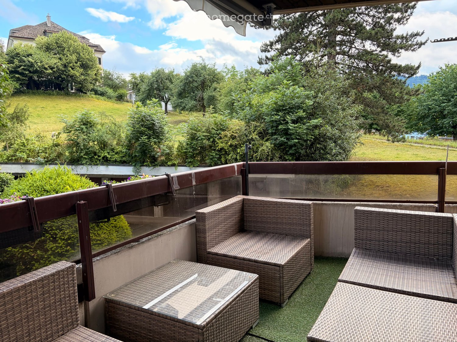 Balcony with wicker furniture, including a sofa and a table, overlooking a green landscape with trees and a house in the background.