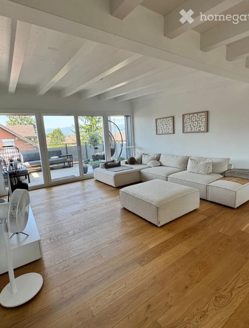 Modern living room with light-colored sectional sofa, ottoman, and a white fan. Large windows offer a scenic view of the outdoors.