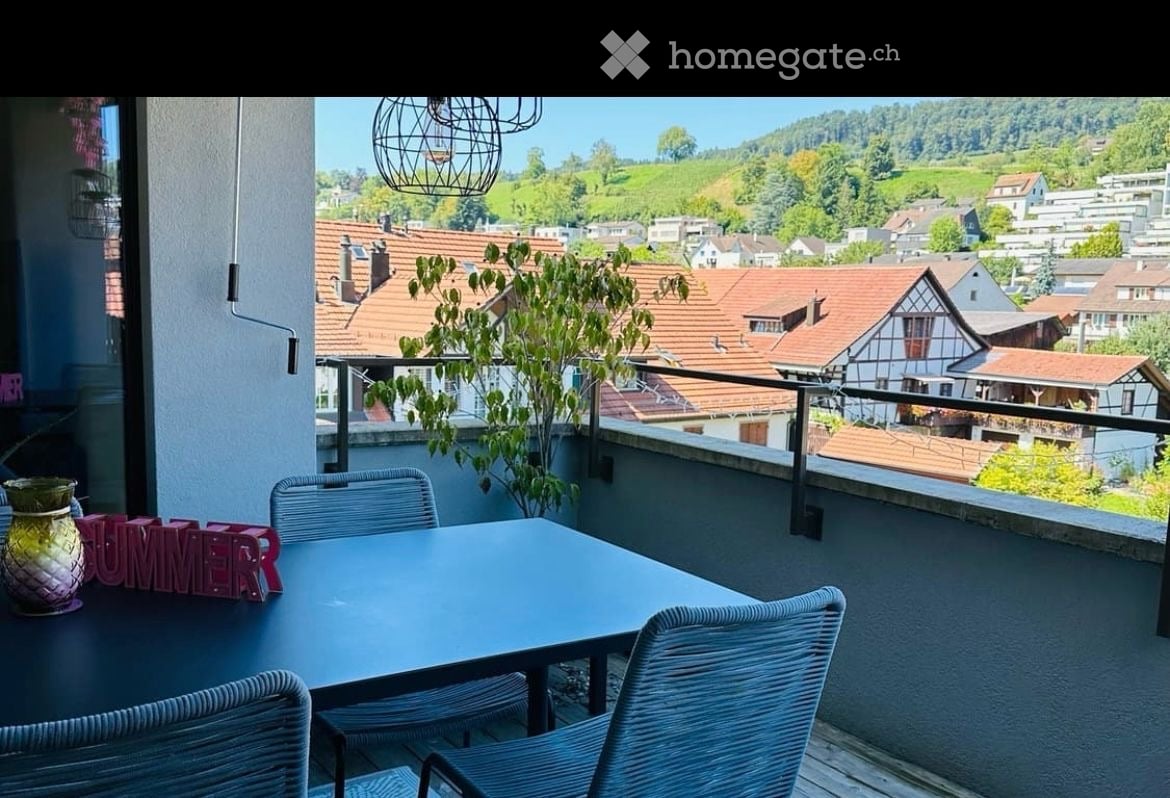 Balcony with a scenic view of a town and hills, featuring a table, chairs, and a decorative vase.