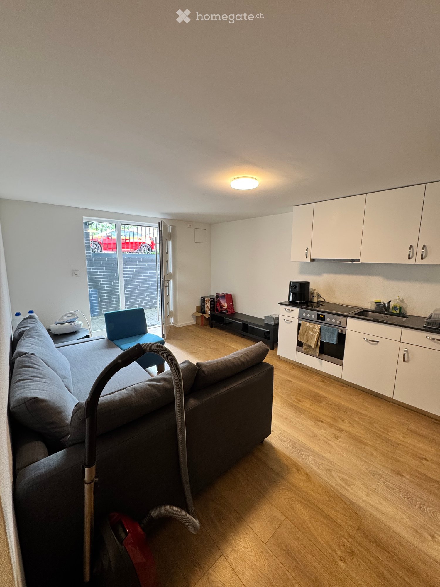 A modern, open-plan living and dining area with a grey sofa, a blue chair, a coffee table, and a small kitchen area with white cabinets, an oven, and a sink.