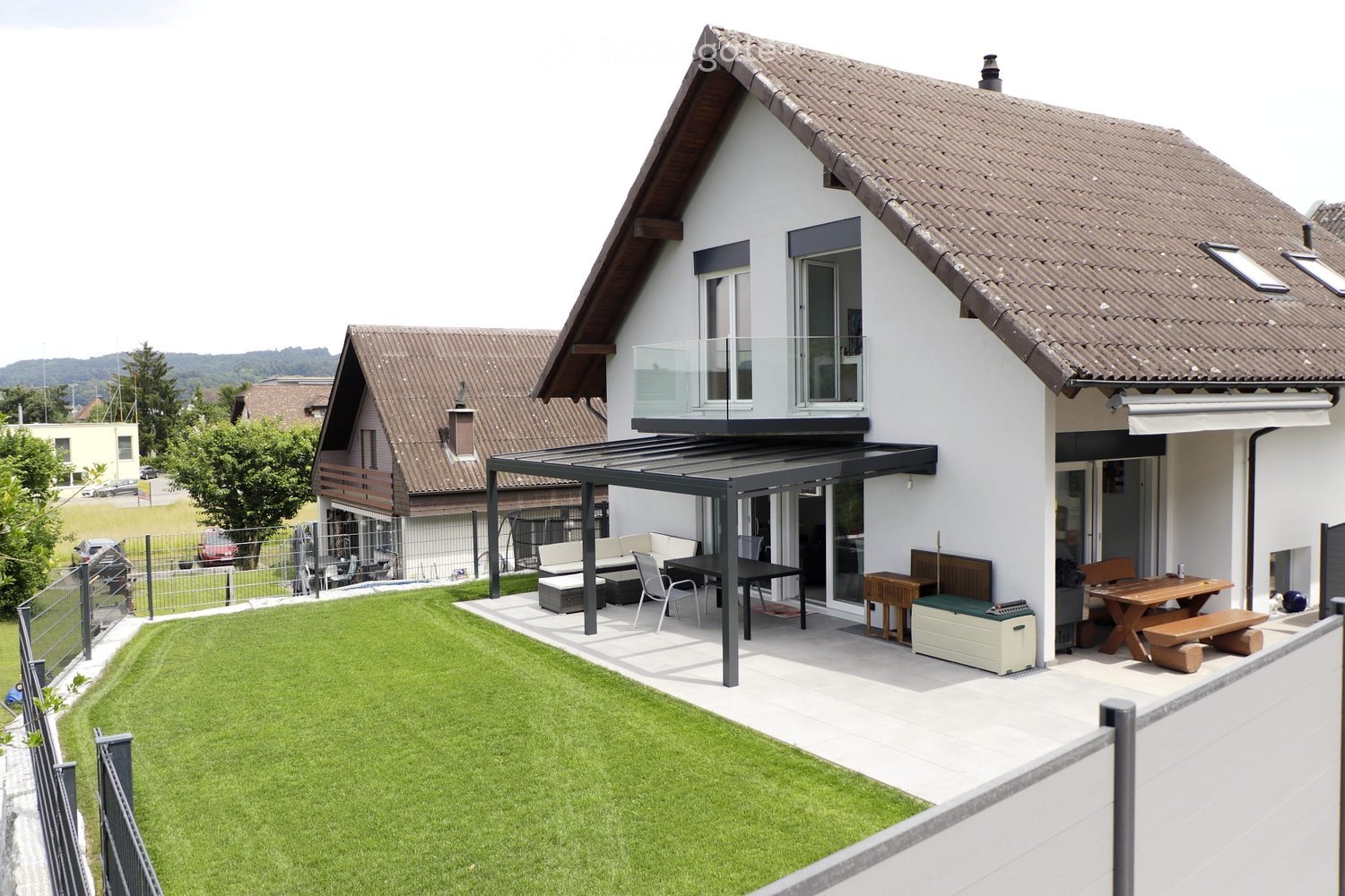 Modern two-story house with a well-maintained lawn, patio area, and outdoor furniture including a sofa, table, chairs, and a picnic table.
