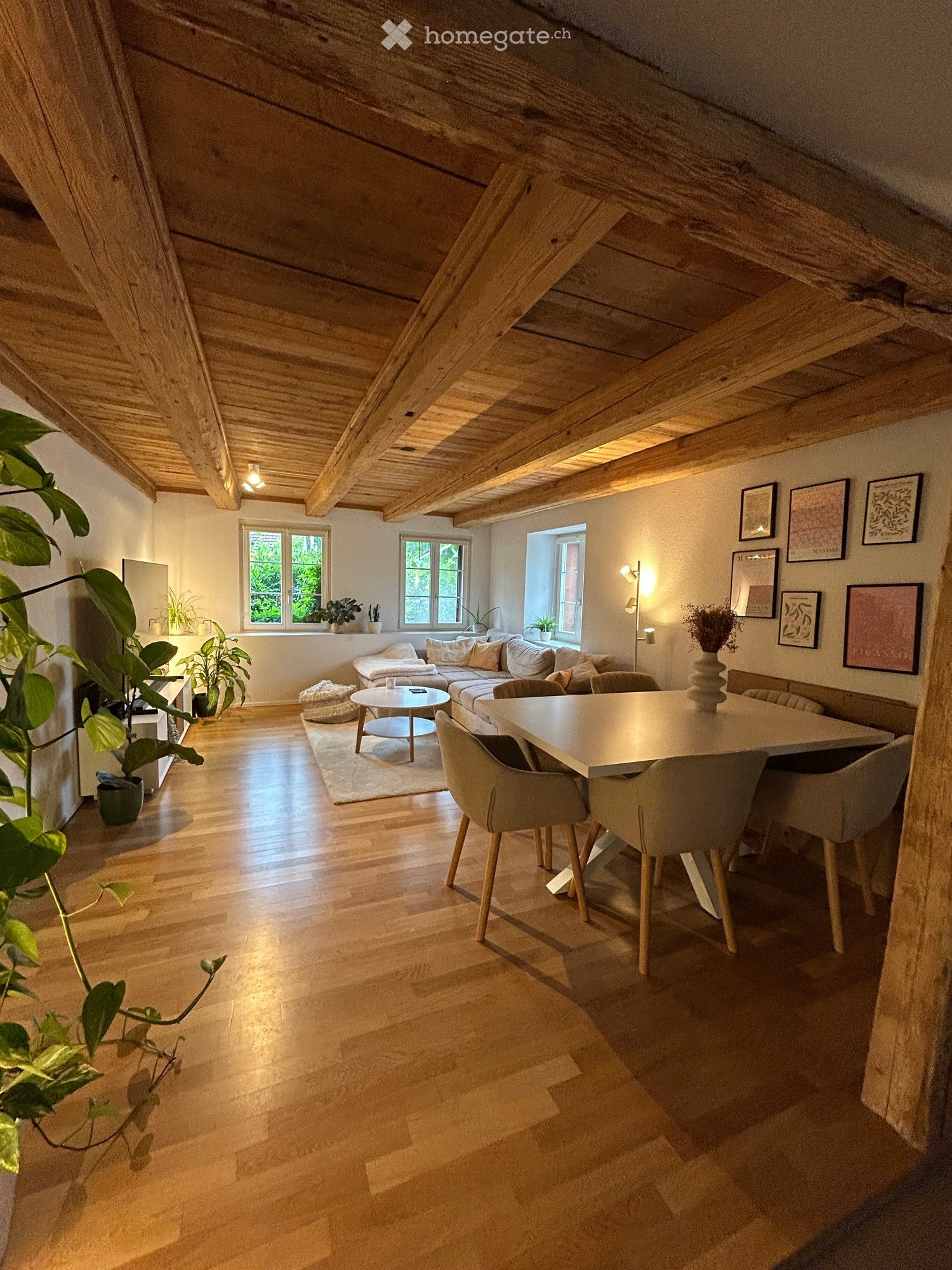 Open-concept living and dining area with exposed wooden beams, hardwood flooring, large windows, sofa, coffee table, dining table, and chairs.