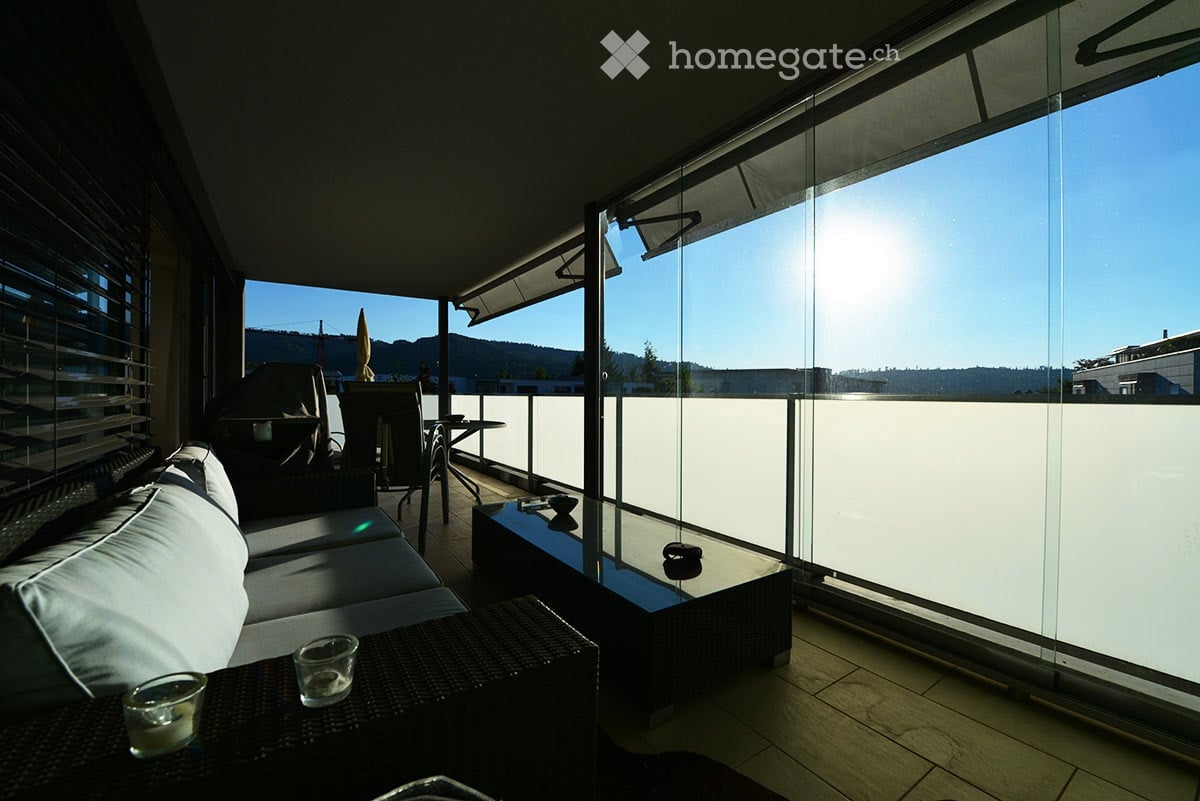 Modern balcony with large glass windows, sofa, coffee table, chairs, and umbrella, overlooking a scenic view.