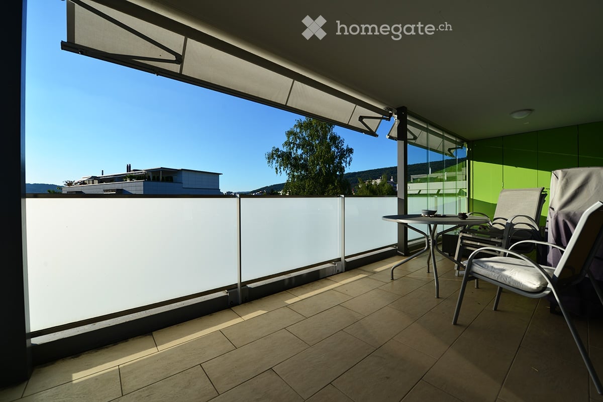 Balcony with tiled floor, white railing, and outdoor furniture including a table and chairs, offering a view of the surrounding area.