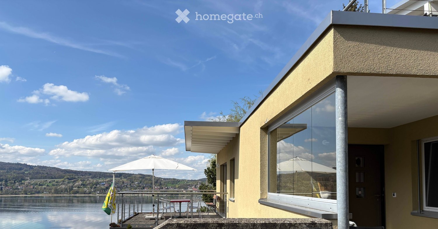 Balcony with a scenic view of a lake and surrounding landscape, featuring a table, chairs, and an umbrella.