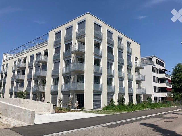 Modern multi-story building with balconies, wide facade, concrete sidewalk, paved street, green lawn, nearby road