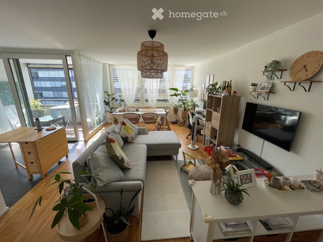 Modern living room with a grey sectional sofa, coffee table, dining area with chairs and table, large windows with sheer curtains, wall-mounted TV, shelves with decorative items, and various plants.