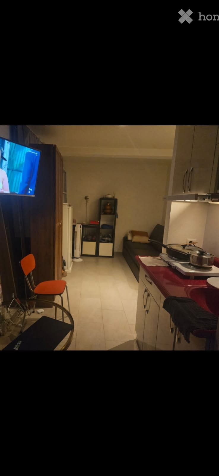 A compact, multi-functional room with a small kitchen area, a couch, a television, and a chair. The kitchen features a counter, sink, and cabinets, while the living area includes a couch and a television.