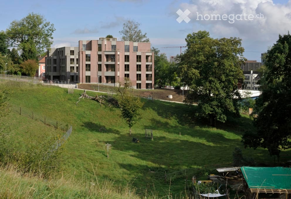 Modern multi-story residential building with a raised ground floor, surrounded by green lawn and trees.