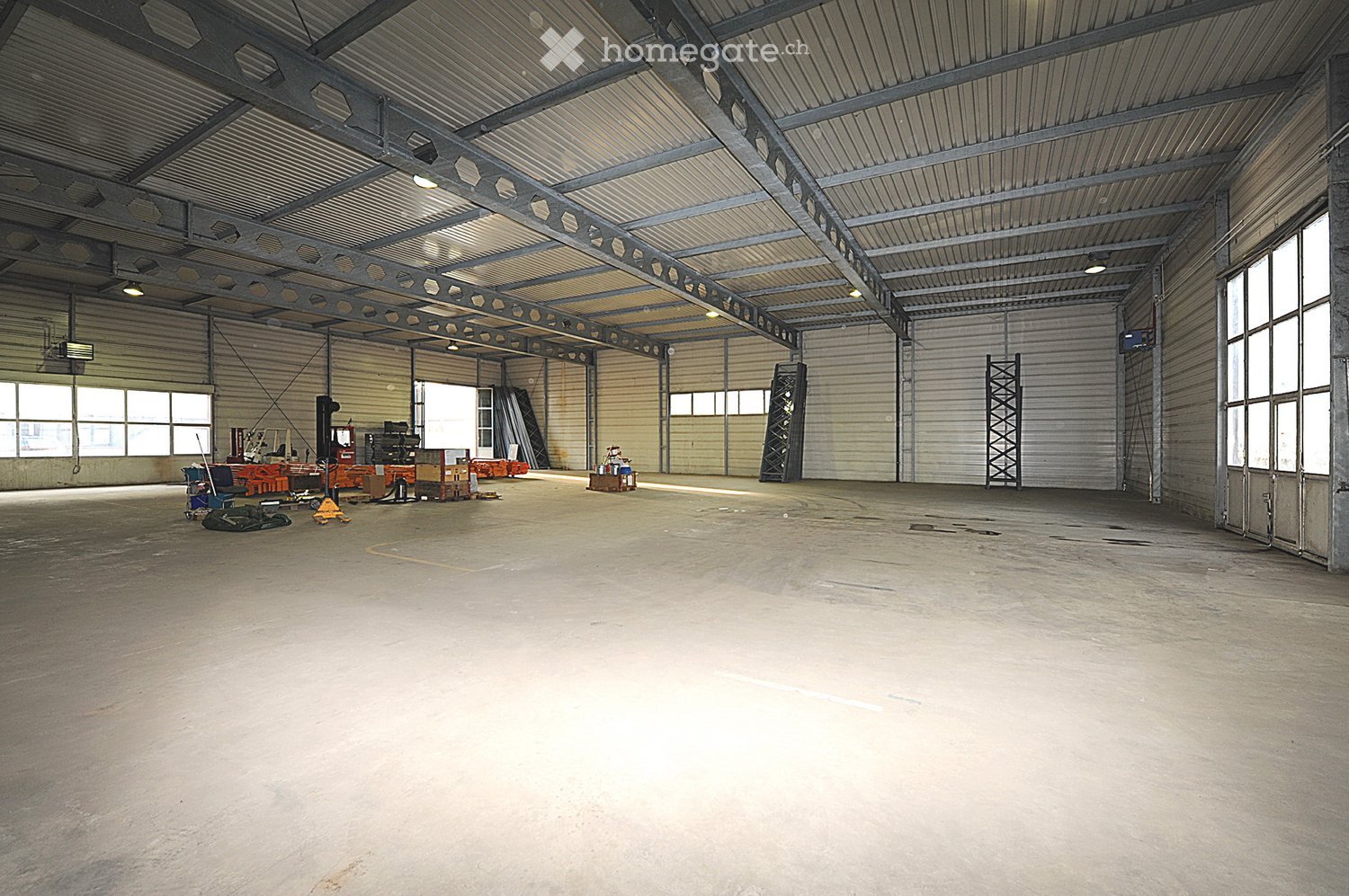 Large, empty industrial-style space with a high, corrugated metal ceiling and exposed beams. The floor is concrete, and the walls are metal panels with large windows allowing natural light. There are no furnishings or equipment visible.