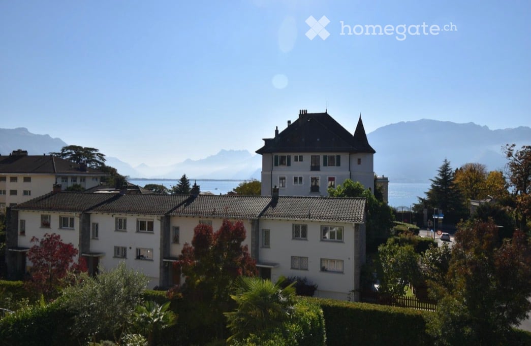 A multi-story residential building with a classic architectural style, surrounded by greenery and offering a scenic view of mountains and a body of water in the background.