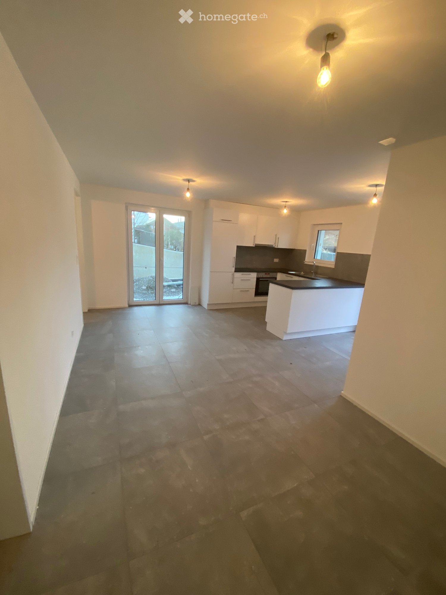 Empty room with tiled floor, white walls, and a kitchen area with white cabinets and a countertop. There is a sliding glass door leading outside.