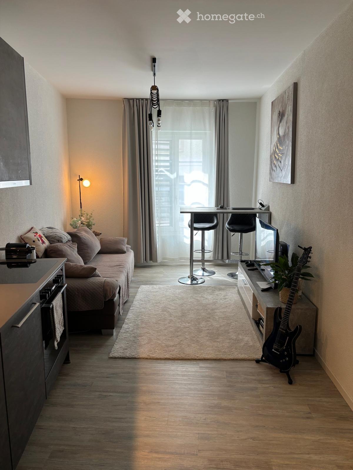 A modern, compact living space with a sofa, a small table with two stools, a guitar, a laptop, and a plant. The room features wooden flooring, a large window with curtains, and a painting on the wall.