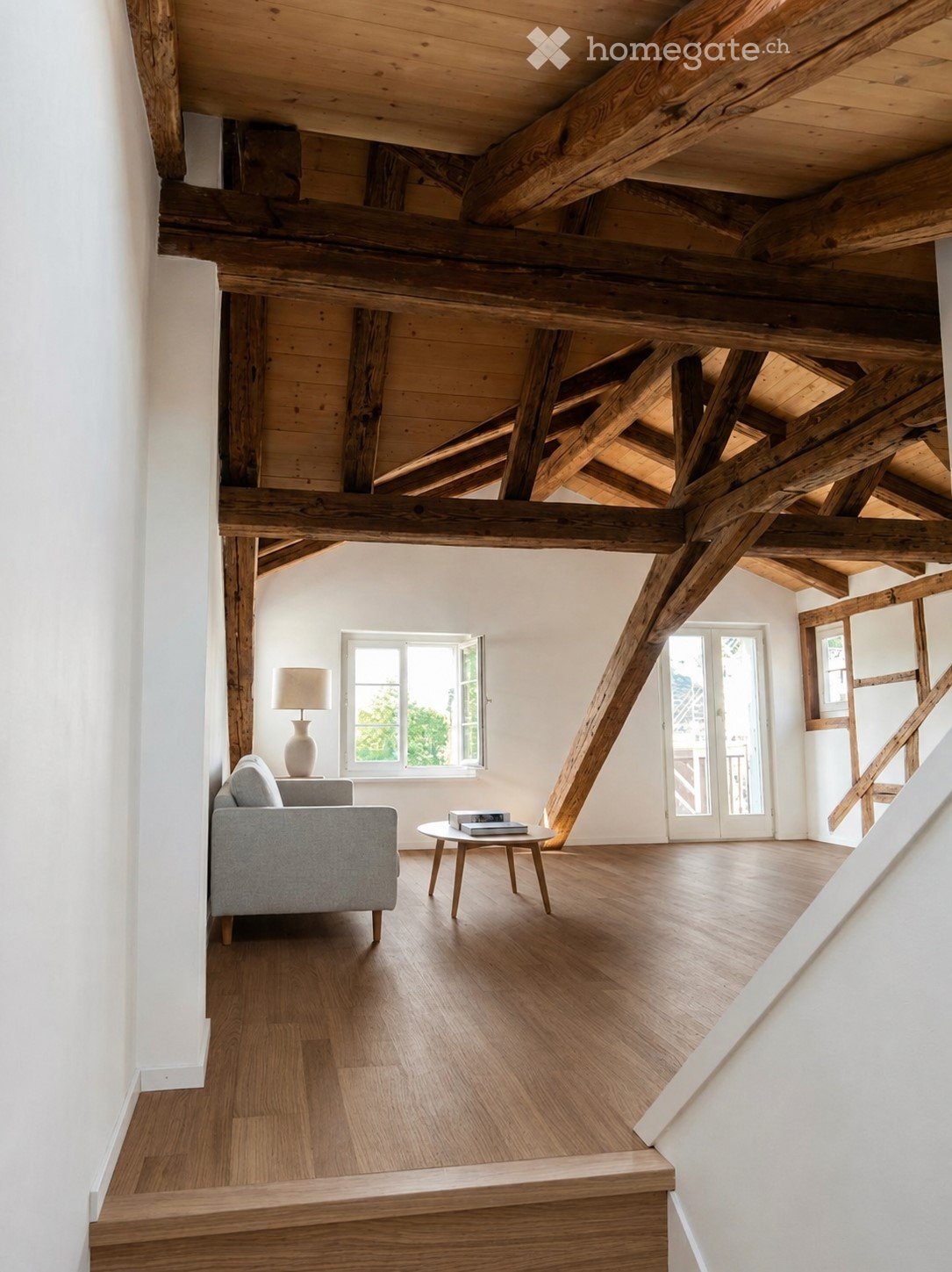 Loft-style living room with exposed wooden beams, hardwood flooring, a grey sofa, a small round coffee table, and a table lamp.