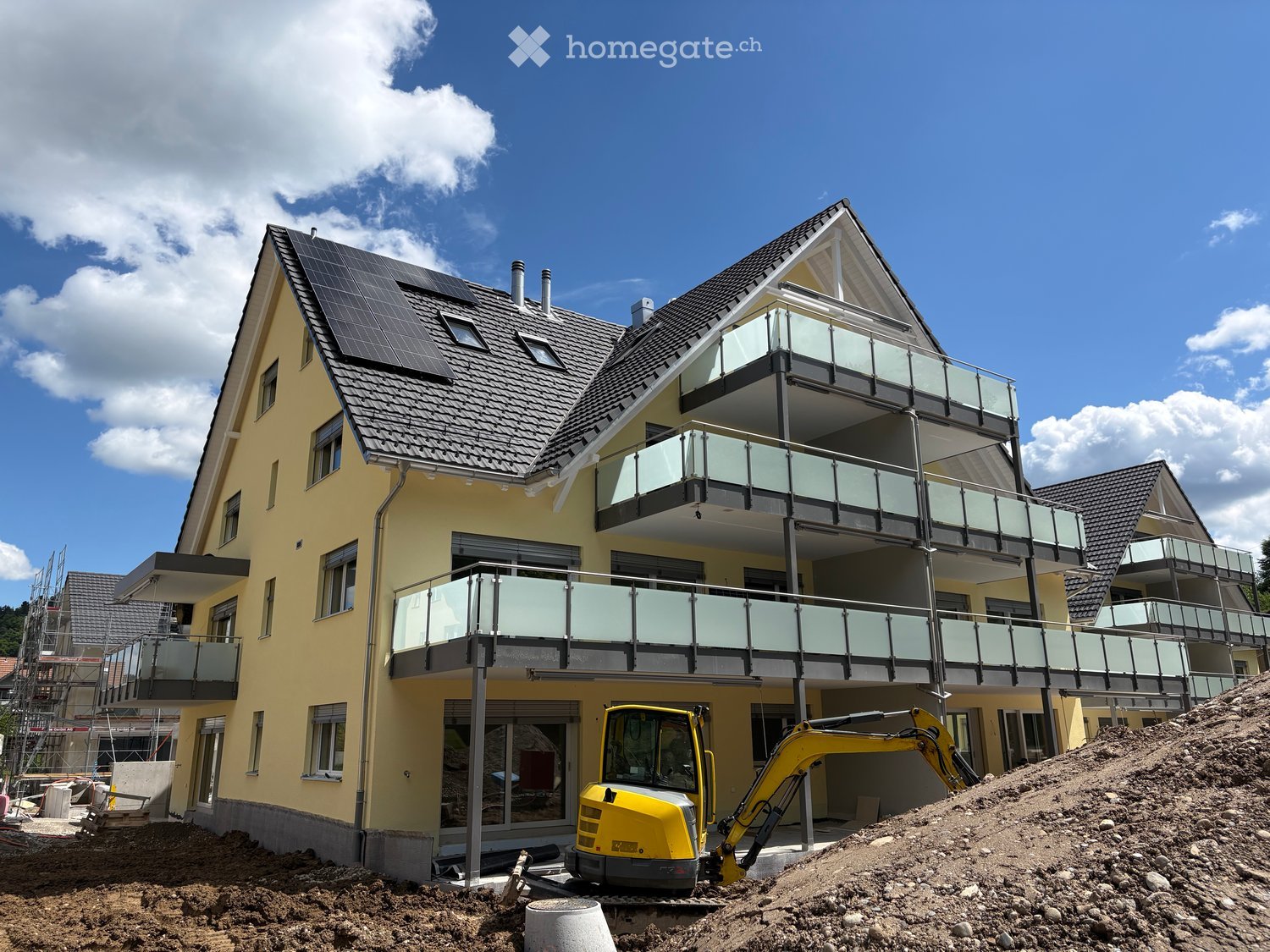 Modern two-story house with yellow exterior, multiple balconies, solar panels on the roof, and construction equipment nearby.