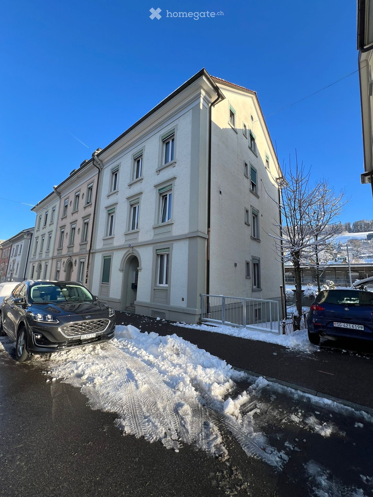 A multi-story residential building with white facade and multiple windows, parked cars in front, and snow on the ground.