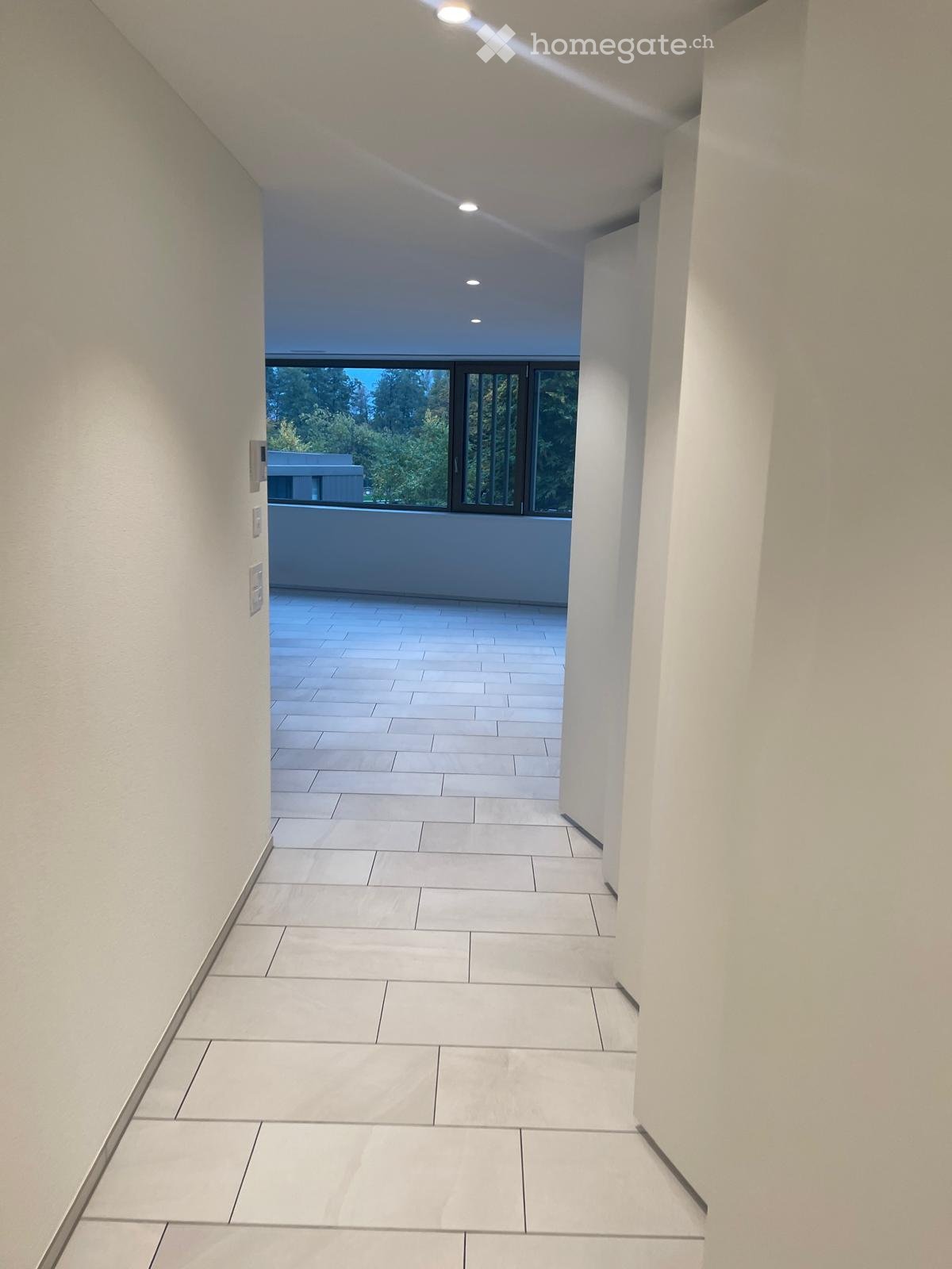 Modern hallway with white tiled flooring, white walls, and recessed lighting. The hallway leads to a balcony area with large windows offering a view of greenery outside.