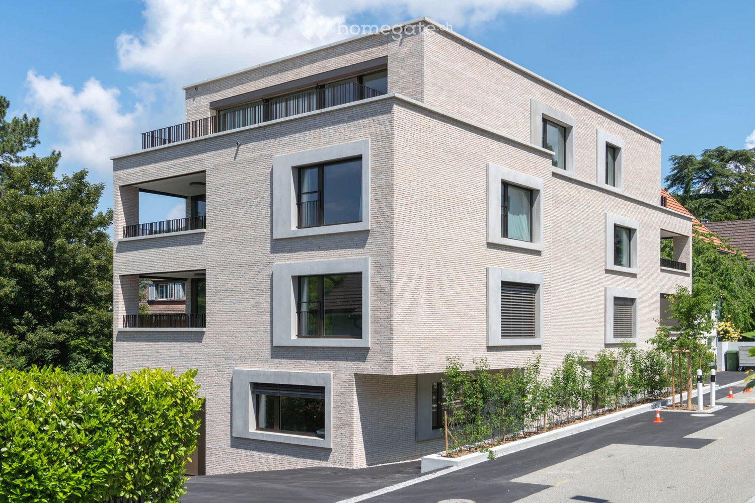 Modern three-story building with light gray brick facade, large windows, and balconies. The structure features clean lines and contemporary design, surrounded by greenery and a paved driveway.