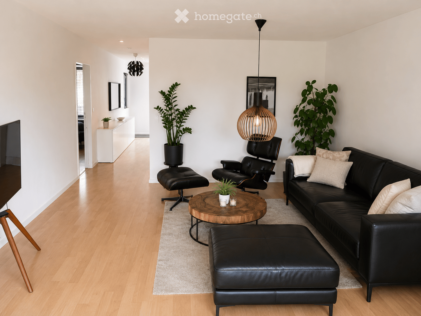 Modern living room with light wooden flooring, white walls, and large windows. Features a black leather sofa with white cushions, a matching ottoman, a round wooden coffee table, a black leather armchair, and a hanging pendant light. Two large potted plan