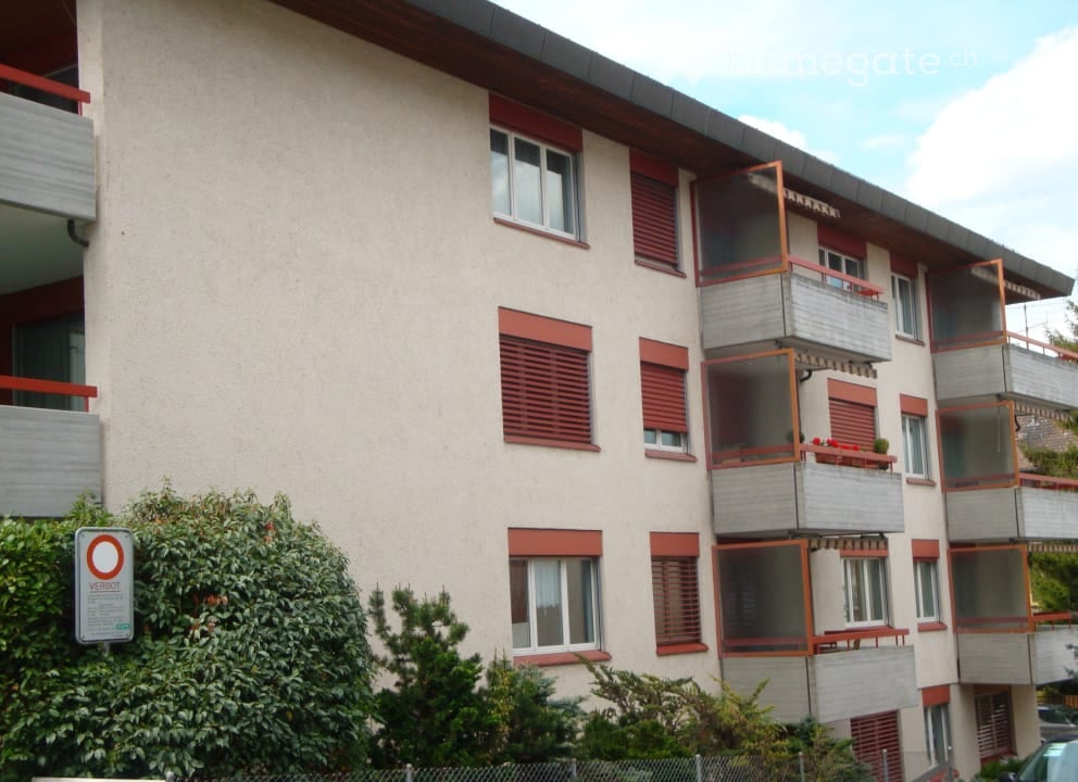 Four-story residential building with beige facade, red shutters, and multiple balconies with railings.