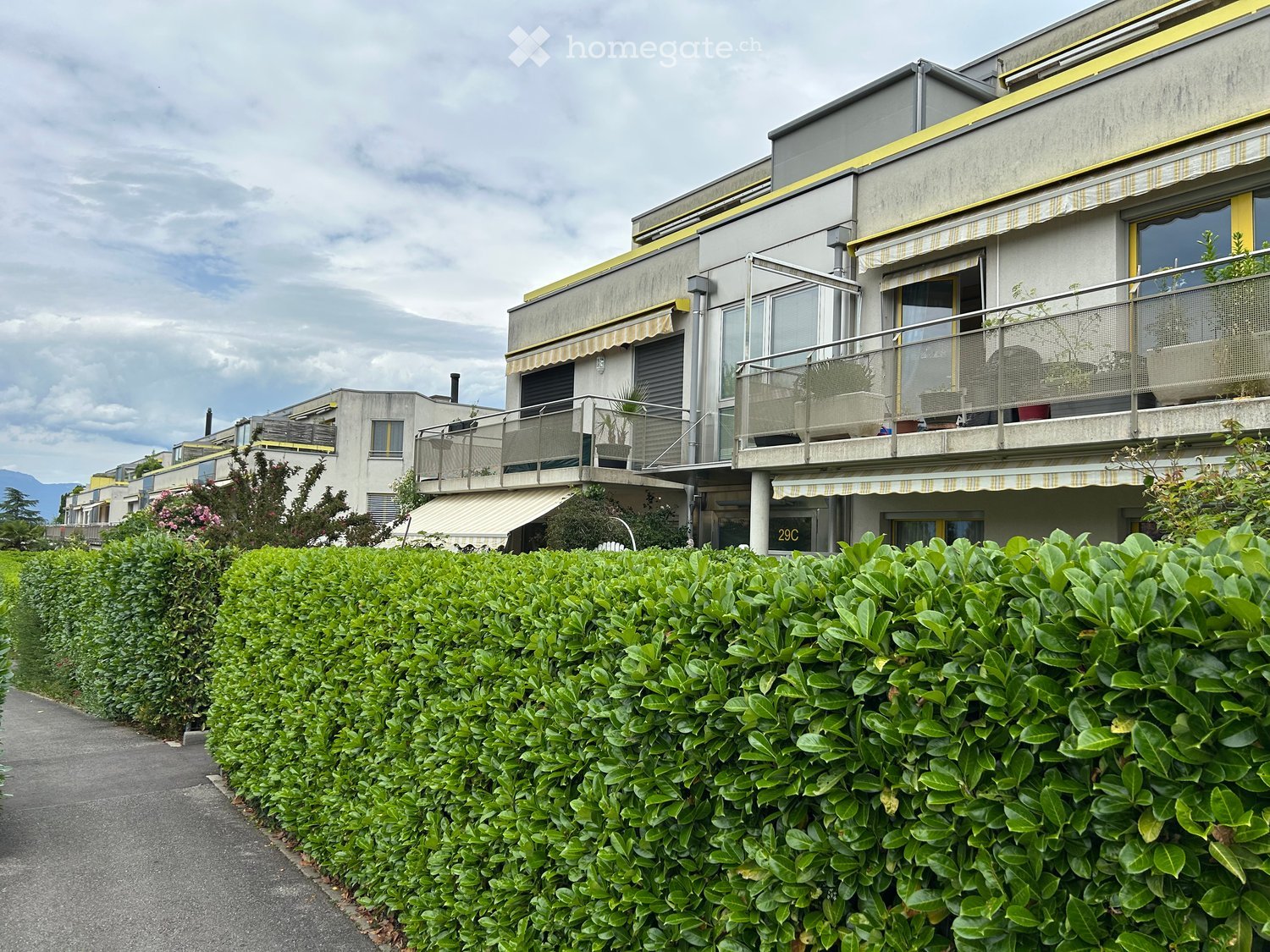 Modern multi-story building with balconies, large windows, and a neatly trimmed hedge in front.