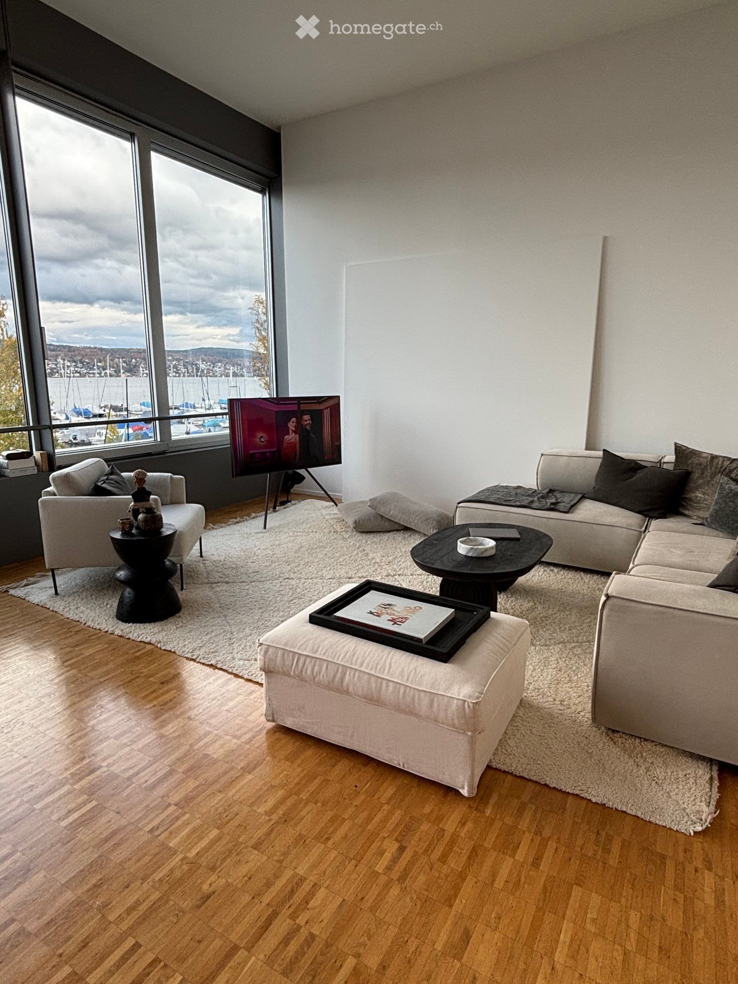 Modern living room with large windows offering a view of the harbor, featuring a sectional sofa, coffee table, ottoman, armchair, and a flat-screen TV on a stand.